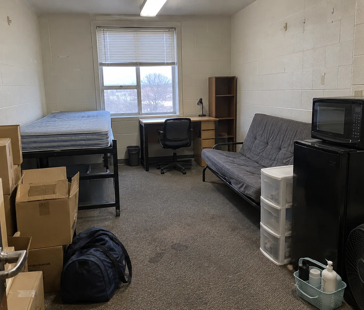 Dorm room at move-out with a stripped mattress, futon, mini fridge and boxes packed by the door