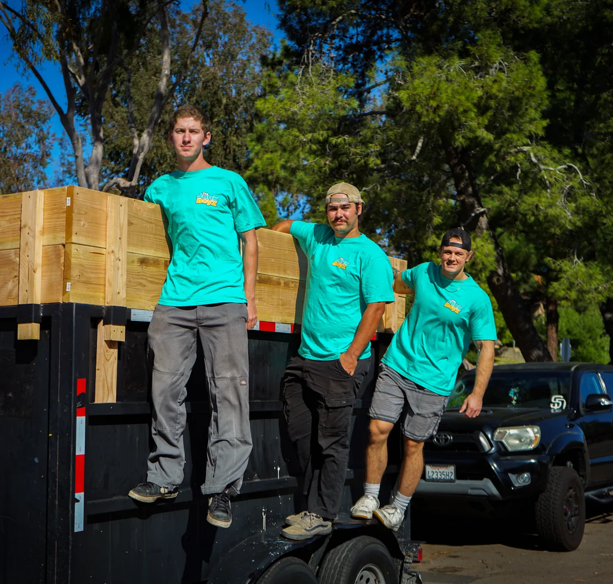 Three Junk Boyz crew members in teal shirts standing on the side of the dump trailer